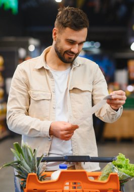 Gülümseyen, uzun basılı bir faturaya bakan, süpermarket koridorlarındaki harcamaları ve alışverişleri analiz eden bir adam.