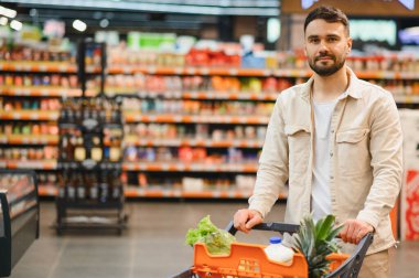 Genç adam market alışverişi yapıyor, içi taze yiyecek ve malzemelerle dolu bir yük arabasını parlak bir süpermarkette itiyor.