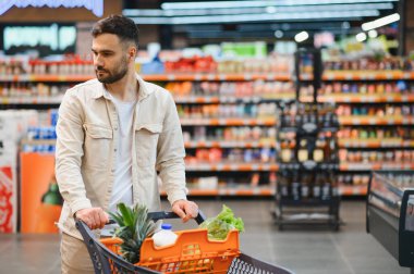 Market alışverişi yapan genç bir adam perakende mağazasının kalabalık koridorlarında dolu bir sepeti itiyor. Tüketim kavramı