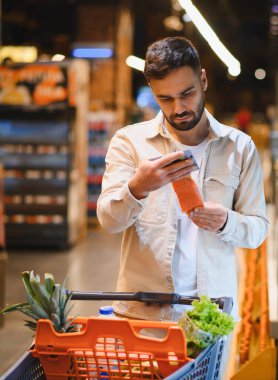 Market koridorunda somon filetosunu taramak ya da araştırmak için akıllı telefonunu kullanan bir adam.