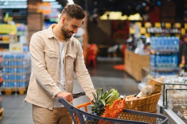 Taze ürünlerle dolu bir market arabasını iten, modern bir süpermarketten sağlıklı şeyler alan mutlu bir adam.