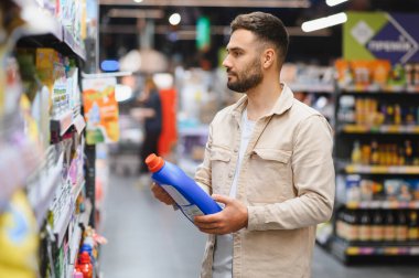 Müşteri elinde bir şişe temizlik malzemesi tutarken modern bir markette raflardaki ev eşyalarına göz atıyor.