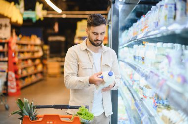Elinde bir şişe süt tutan, marketin mandıra bölümünde alışveriş yaparken etiketi okuyan bir adam.