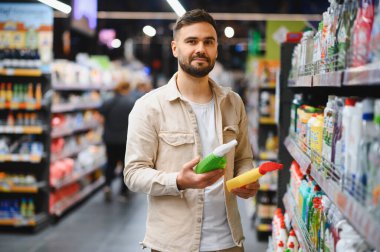 Parlak bir süpermarket koridorunda iki şişe temizlik malzemesini karşılaştıran bir adam hangi ev ürününü alacağına karar verir.