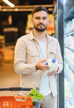 Süpermarkette süt satan genç bir adam, içi taze ürünlerle dolu bir alışveriş arabasını itiyor.