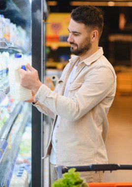 Süpermarkette soğutulmuş bir raftan süt alan bir adam. Elinde bir karton var ve ürünü kontrol ediyor.