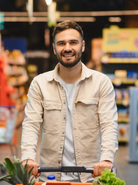 Gülümseyen, alışveriş arabasını iten genç bir adam. Parlak bir perakende mağazası koridorunda içi taze yiyeceklerle dolu.