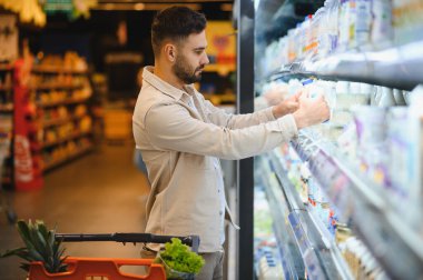 Genç adam markette alışveriş yapıyor, süt ürünlerine bakıyor ve bir seçim yapmaya karar veriyor, yakınlarda tam bir alışveriş arabası.