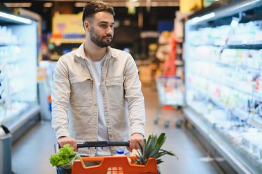 Bir süpermarket koridorunda alışveriş arabasını iten genç adam taze ürünler ve günlük ihtiyaçlar seçiyor, seçeneklere odaklanıyor.