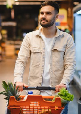 Market alışverişi yapan genç bir adam, süpermarket koridorunda dolu bir arabayı iterken, seçilmiş eşyaları seçmeye odaklanmış görünüyor.