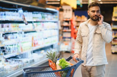 Markette cep telefonuyla konuşurken market arabalarını iten genç bir adam.