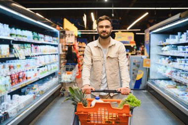 Market koridorunda içi taze ürünlerle dolu turuncu bir arabayı iten genç adam.