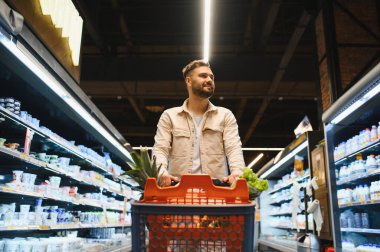 Parlak bir süpermarket koridorunda yiyecek alışverişi yapan bir adam taze ürünler ve bakkaliyelerle dolu bir arabayı itiyor.