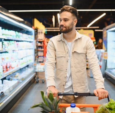 Genç bir adam, parlak bir süpermarket koridorunda içi taze yiyeceklerle dolu bir alışveriş arabasını itip ürün arıyor.