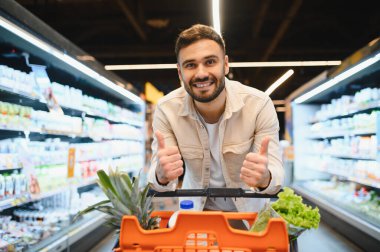 Gülümseyen adam market alışverişinden zevk alıyor, taze ürünlerle dolu bir arabayı itiyor ve olumlu bir jest yapıyor.