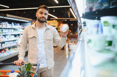 Markette elinde beyaz yoğurt olan bir adam dondurulmuş süt bölümünde alışveriş arabasıyla seçenekleri değerlendiriyor.