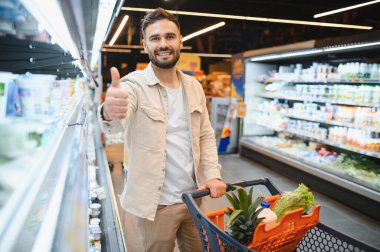 Mutlu müşteri market koridorunda duruyor. Taze yiyecek dolu alışveriş arabalarını itiyor. Memnuniyetini dile getiriyor.