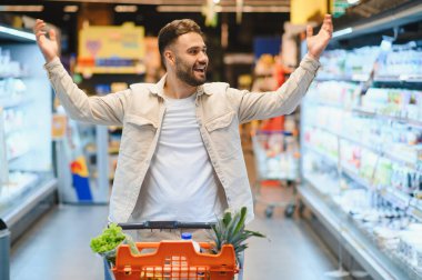 Market koridorunda elinde alışveriş arabasıyla duran sakallı adam. Kolları mutlu bir şekilde kaldırıldı.