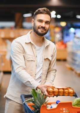 Gülümseyen adam alışveriş arabasını itiyor, yumurta tutuyor ve süpermarketten yiyecek alırken kameraya bakıyor.