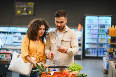 Alışveriş listesine bakan genç bir çift bir süpermarketin koridorlarında içi taze ürünlerle dolu bir arabayı itiyor.