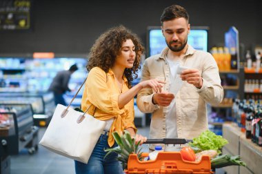 Alışveriş listelerindeki ürünleri uzun bir makbuzla karşılaştıran genç bir çift, modern bir markette bütçe ve harcamaları yönetiyor.