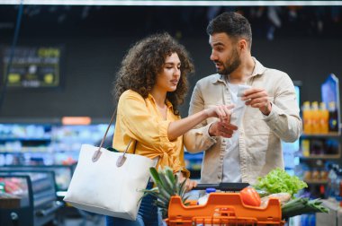 Market fiyatlarını karşılaştıran ve alışveriş listelerini süpermarkette makbuz kullanarak tartışan genç bir çift.
