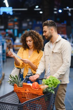 Genç bir çift alışveriş listesine bakıyor ve modern bir süpermarket koridorunda içi taze yiyecek dolu bir arabayı itiyor.