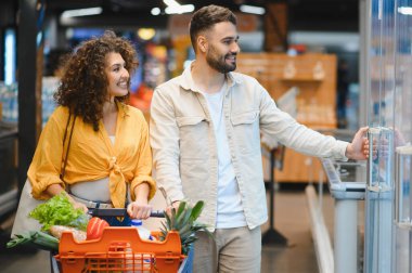 Parlak bir süpermarket koridorunda içi taze meyve ve sağlıklı yiyeceklerle dolu bir market arabasını iten gülümseyen genç çift.