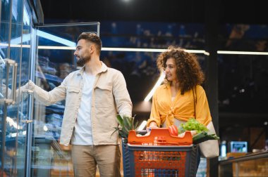 Genç çift, modern bir markette dondurucudan dondurulmuş ürünleri seçiyor ve taze ürünlerle dolu bir arabayı itiyor.