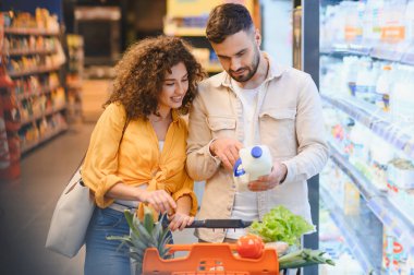 Kalabalık bir süpermarkette yiyecek alışverişi yapan ve ürünleri karşılaştıran genç bir çift.