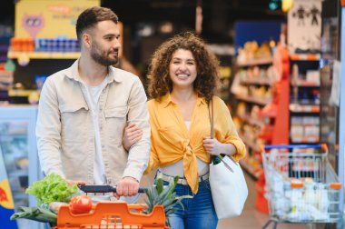Gülümseyen genç çift alışveriş arabalarını itiyor. Taze ürünlerle dolu, günlük market alışverişlerinin keyfini çıkarıyorlar.