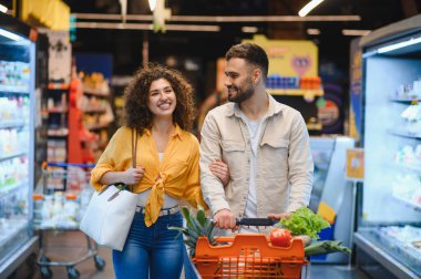 Genç gülümseyen adam ve kadın taze ürünleri seçiyorlar ve modern bir markette alışveriş arabasını koridorda itiyorlar.