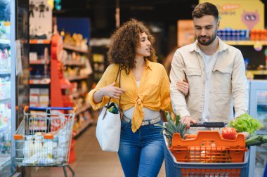 Genç çift alışverişleri tartışırken, parlak ışıklı bir süpermarkette içi taze ürünlerle dolu bir alışveriş arabasını itiyorlar.