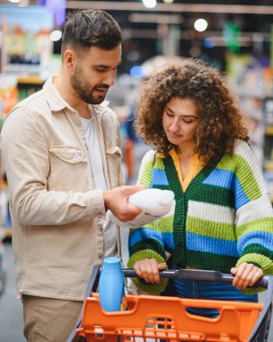 Market alışverişi sırasında ürün etiketini tartışan genç çift, perakende satış ortamında ev ihtiyaçları için ürünleri karşılaştırıyor