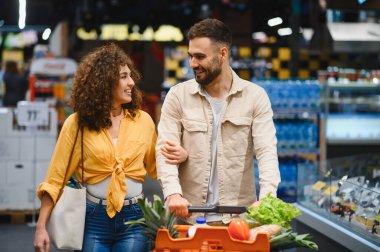 Genç çift birlikte vakit geçiriyorlar, markette yürürken içi taze yiyecek dolu bir arabayı itiyorlar.