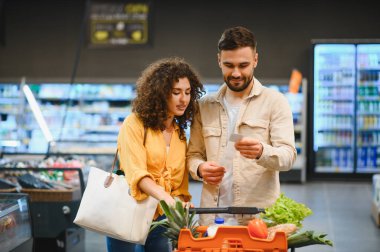 Alışveriş listesine bakan genç bir çift, süpermarkette taze yiyecek ve ürün alırken, alışveriş arabasını itiyor.