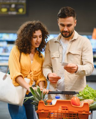 Market listesini ve makbuzu karşılaştıran, alışveriş arabalarını taze yiyeceklerle doldururken bütçeyi yöneten genç çift