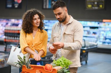 Modern bir süpermarkette alışveriş yaparken alışveriş arabalarındaki ürünlerle listedeki ürünleri karşılaştıran genç bir çift.