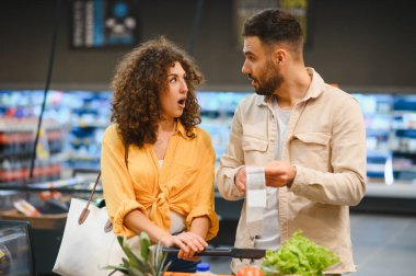 Genç bir çift, gıda fiyatlarının artmasından kaynaklanan ekonomik şoka uğrayan, şaşırmış ifadelerle uzun bir market makbuzuna bakıyor.