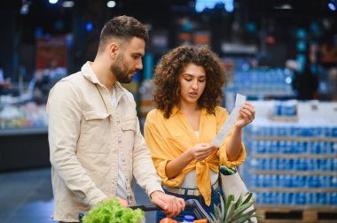 Market fişini alışveriş arabası eşyalarıyla karşılaştıran genç çift, süpermarkette yüksek fiyatlar ve bütçe kaygılarını tartışıyor