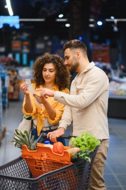 Alışveriş listesindeki ürünlerle market koridorunda yürürken arabalarındaki ürünleri karşılaştıran genç bir çift.