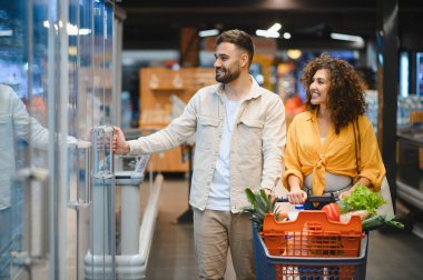 Gülümseyen genç bir çift taze ürünlerle dolu bir alışveriş arabasını itiyor, marketteki soğutuculu vitrinlerden eşyalar seçiyorlar.