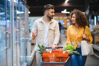 Genç bir çift, modern bir markette içi taze yiyecek dolu bir alışveriş arabasını iterken ürünlerini karşılaştırıyor.