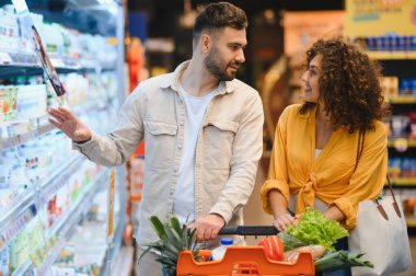 Genç çift soğutuculu bir koridordan ürün seçiyor. Taze ürünler ve temel malzemelerle dolu bir alışveriş arabasını itiyorlar.