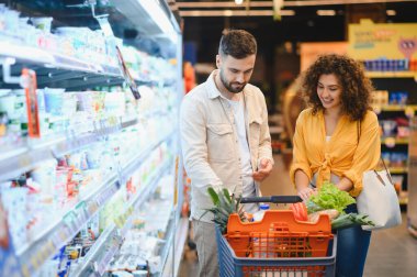 Gülümseyen genç bir çift marketteki soğutuculu raftan ürünleri seçiyor, alışveriş arabalarını taze ürünlerle dolduruyorlar.