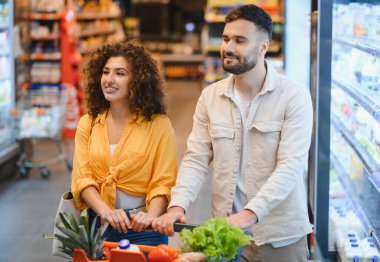 Genç çift gülümsüyor ve alışveriş arabasını itiyor. Modern bir süpermarketin koridorunda içi taze yiyeceklerle dolu.