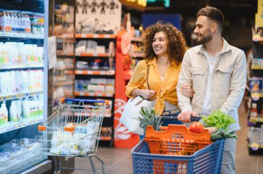 Genç çift gülümserken taze ürünlerle dolu bir arabayı itiyor. Parlak, modern bir süpermarket koridorunda sağlıklı yiyecekler seçiyorlar.