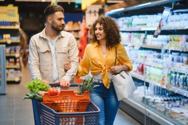 Parlak bir süpermarket koridorunda alışveriş arabasını iterken gülümseyen mutlu çift, taze ürünleri birlikte seçiyorlar.