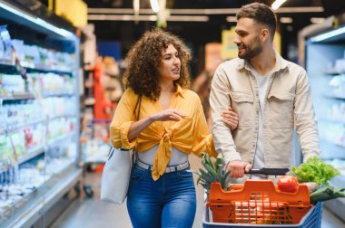 Milenyum çifti yeni ürünler seçiyor ve alışveriş arabasını süpermarket koridorunda itiyorlar.