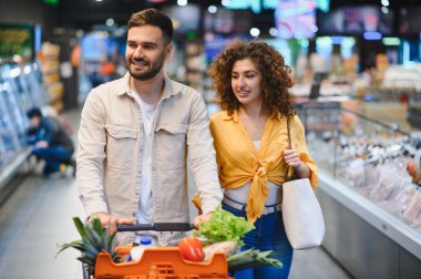Genç bir çift, içi taze ürünlerle dolu bir alışveriş arabasını itiyor. Modern bir market koridorunda eşyalar seçiyorlar.